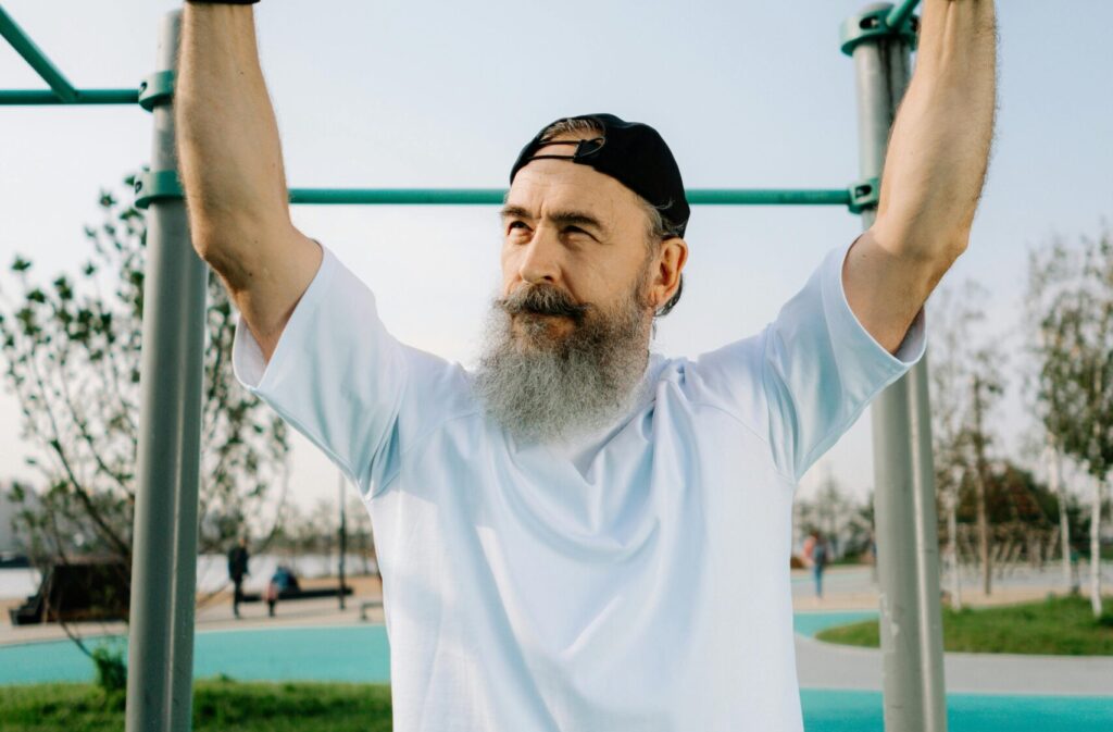 Älterer Mann mit grauem Bart und Cap, der an einer Klimmzugstange trainiert, ein Beispiel für aktiven Muskelaufbau im Alter.
